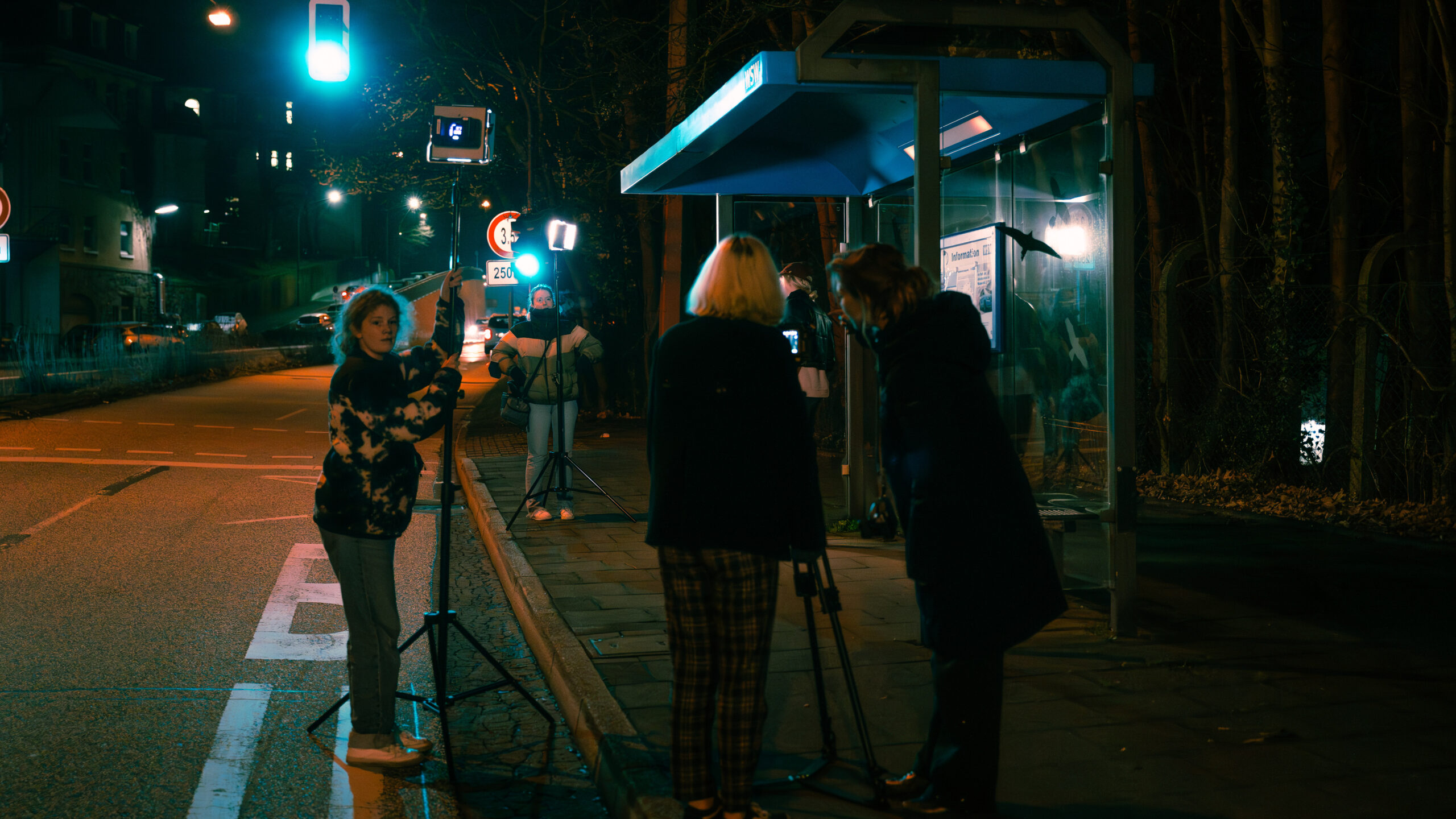 Eine situative Aufnahme hinter den Kulissen eines Filmdrehs an einer nächtlichen Bushaltestelle. Zwei Personen stehen an zwei Lichtpanels, zwei weitere Frauen an einer Stativkamera.