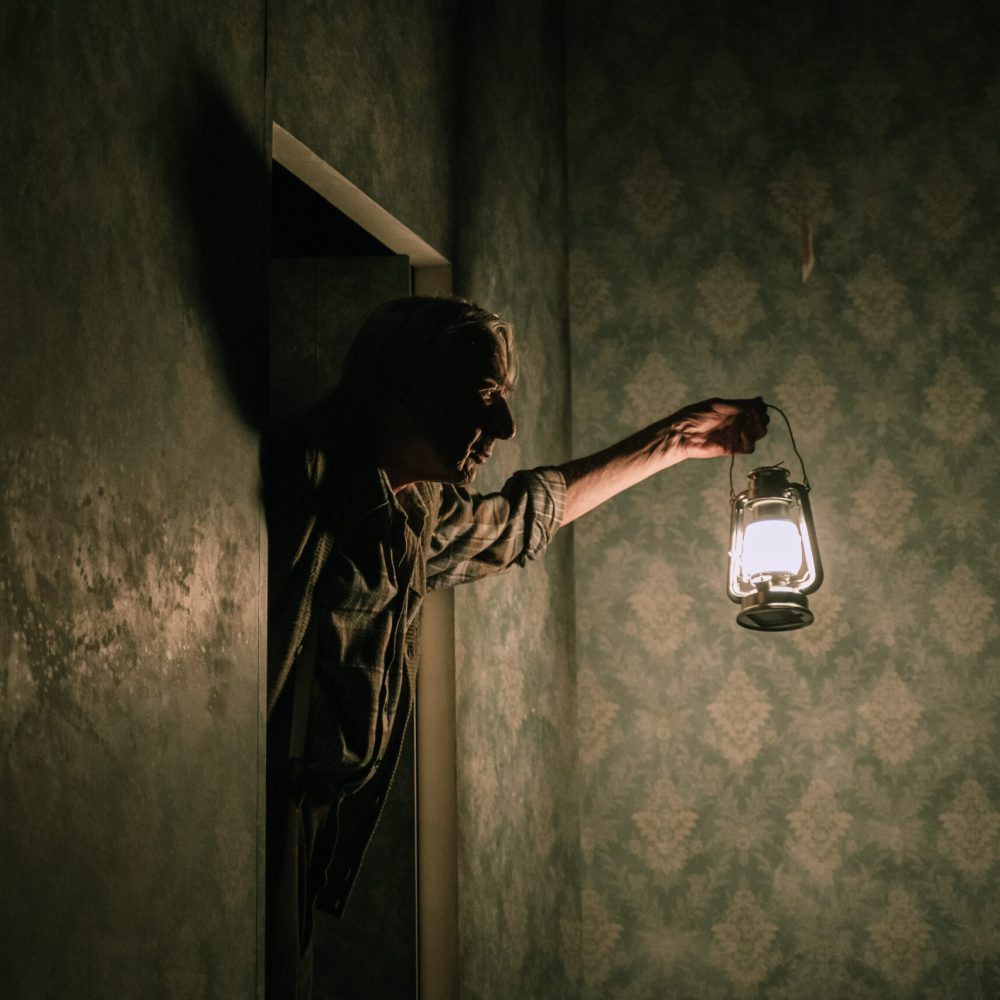 Ein dunkles Bild mit einer Person. Sie schaut durch eine Tür in ein herrschaftliches Zimmer. Der Mann trägt eine Laterne in der Linken Hand, um im Dunkel etwas sehen zu können.